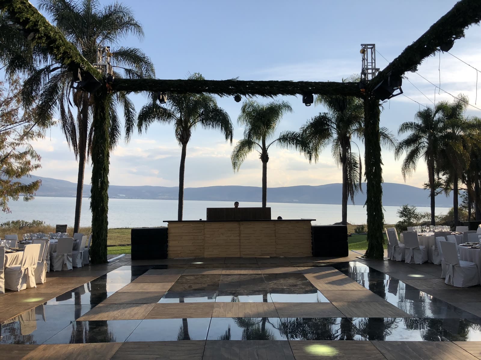 Estructura de gran soporte con follaje enmarcando cabina de DJ y pista espejo frente al lago