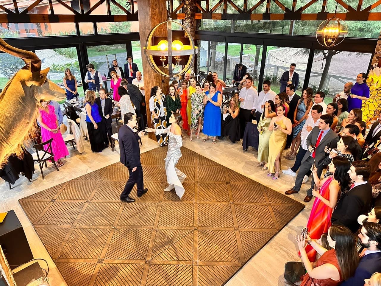 Primer baile de novios sobre pista de madera con patrón geométrico, vista aérea con invitados alrededor
